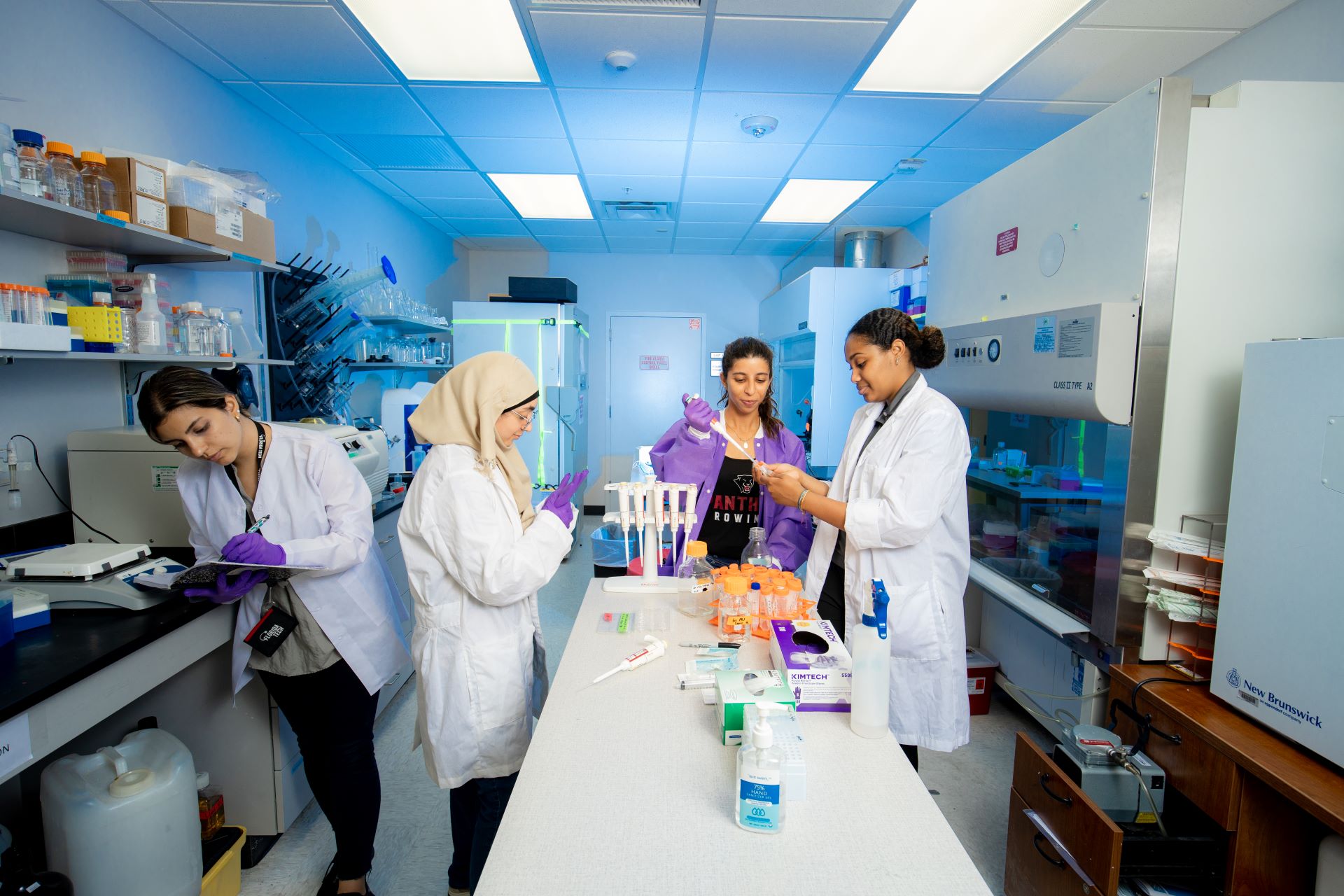 Four female student scientists in lab coats working together in a laboratory, handling test tubes and conducting experiments with various scientific equipment